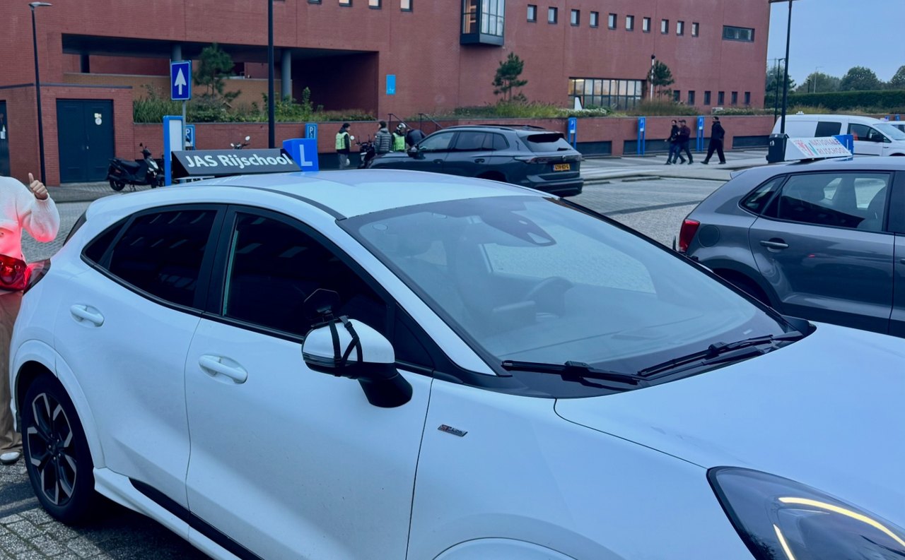 De witte Ford Puma-lesauto van JAS-Rijschool met het blauwe L-bord en het JAS Rijschool-dakbord
